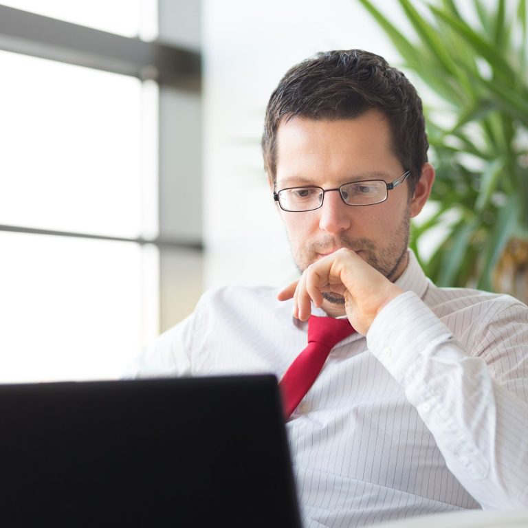 Portrait of successful young businessman in bright modern office focused on work on his laptop computer wearing glasses. Business and entrepreneurship concept.